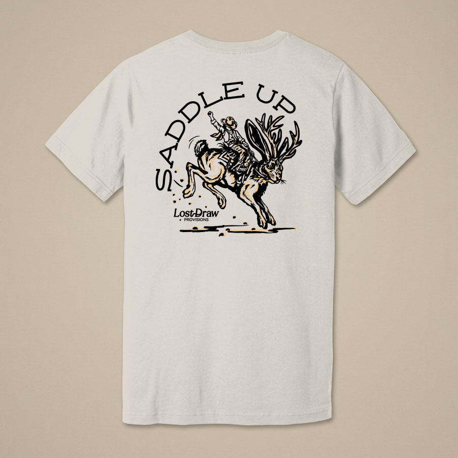A white short-sleeve t-shirt with a black graphic print on the back, featuring the text 'SADDLE UP' and an illustration of a cowboy riding a bucking horse. The Lost Draw Brewing logo is printed below the graphic.