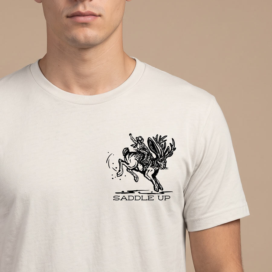 A man wearing a white crewneck t-shirt with a black graphic of a rider on a bucking deer and the text 'SADDLE UP' printed on the chest, set against a solid beige background.