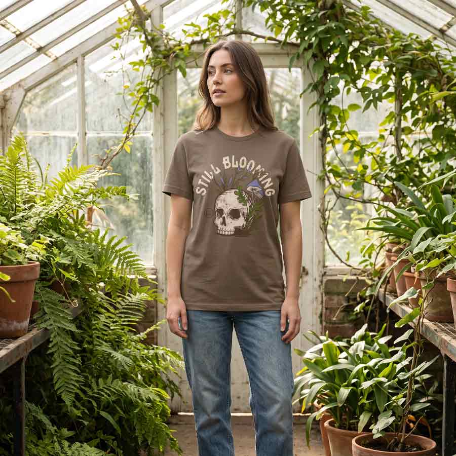 Woman wearing the Still Blooming Jersey Tee inside a greenhouse, displaying the vintage skull and botanical artwork.