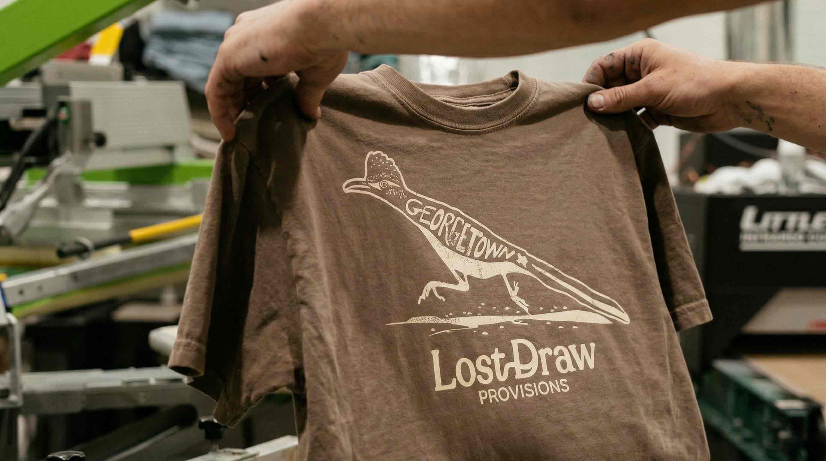 Brown t-shirt with a dinosaur graphic and 'LostDraw' branding held by a person in an industrial setting.