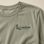Details of Lost Hawk Jersey Tee in Thyme green featuring a small forest-green Lost Draw Provisions hawk logo on the left chest.
