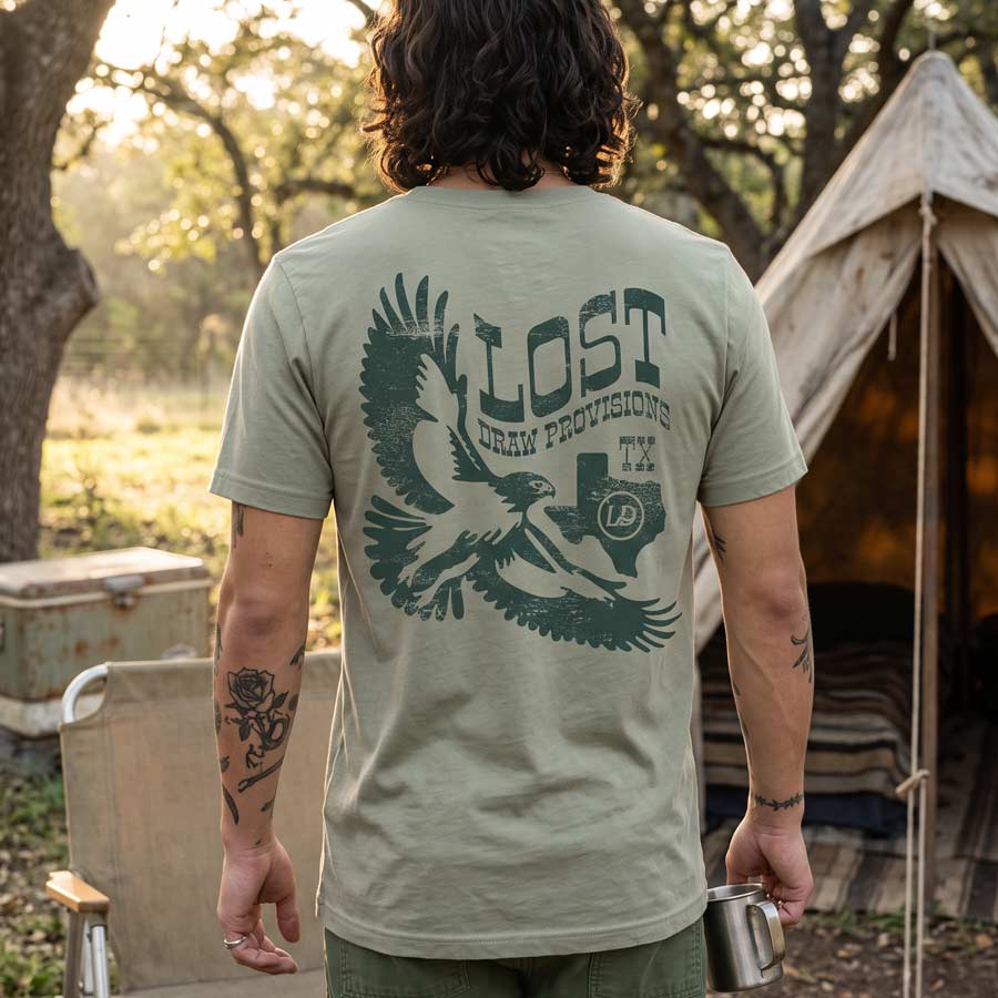 Lifestyle image of back of the Lost Hawk Jersey Tee featuring distressed forest-green hawk illustrations, vintage Lost Draw Provisions lettering, and Texas artwork on a Thyme green shirt.
