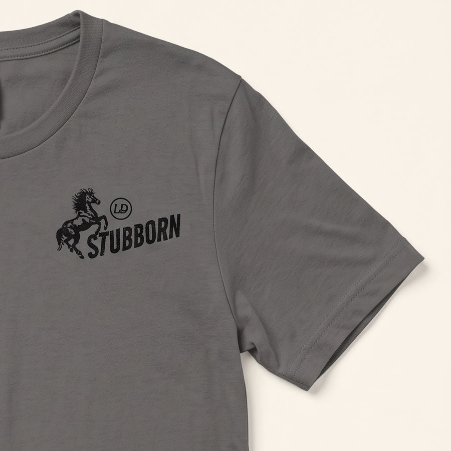 A close-up of a dark gray crewneck t-shirt featuring a black graphic print on the left chest with a rearing horse, a circular 'LP' logo, and the word 'STUBBORN'.