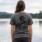 Woman wearing the Too Stubborn Tee beside a freshwater lake, displaying the distressed horse and Too Stubborn to Break back graphic.