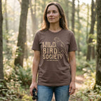 Woman wearing the Wild Bird Society Tee on a woodland trail, displaying the vintage bird illustration and cream lettering.