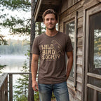 Man wearing the Cocoa Wild Bird Society Tee outside a lakeside cabin, showing the distressed bird graphic.