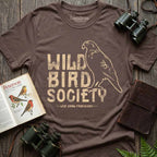 Cocoa Wild Bird Society Tee displayed with binoculars, a bird field guide, and a notebook, featuring the vintage cream bird graphic.